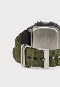 Montre numérique avec un boîtier noir, un dos en acier inoxydable et un bracelet en tissu vert. Elle dispose d'une boucle en argent et de plusieurs trous d'ajustement.