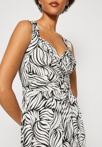 Femme portant une robe sans manches à imprimé zèbre noir et blanc, avec taille froncée et détail anneau, montrant partiellement le haut du corps et le visage.