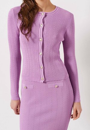Femme portant un cardigan en maille côtelée lavande ajusté avec des boutons en perles et une jupe assortie avec des poches avant boutonnées.