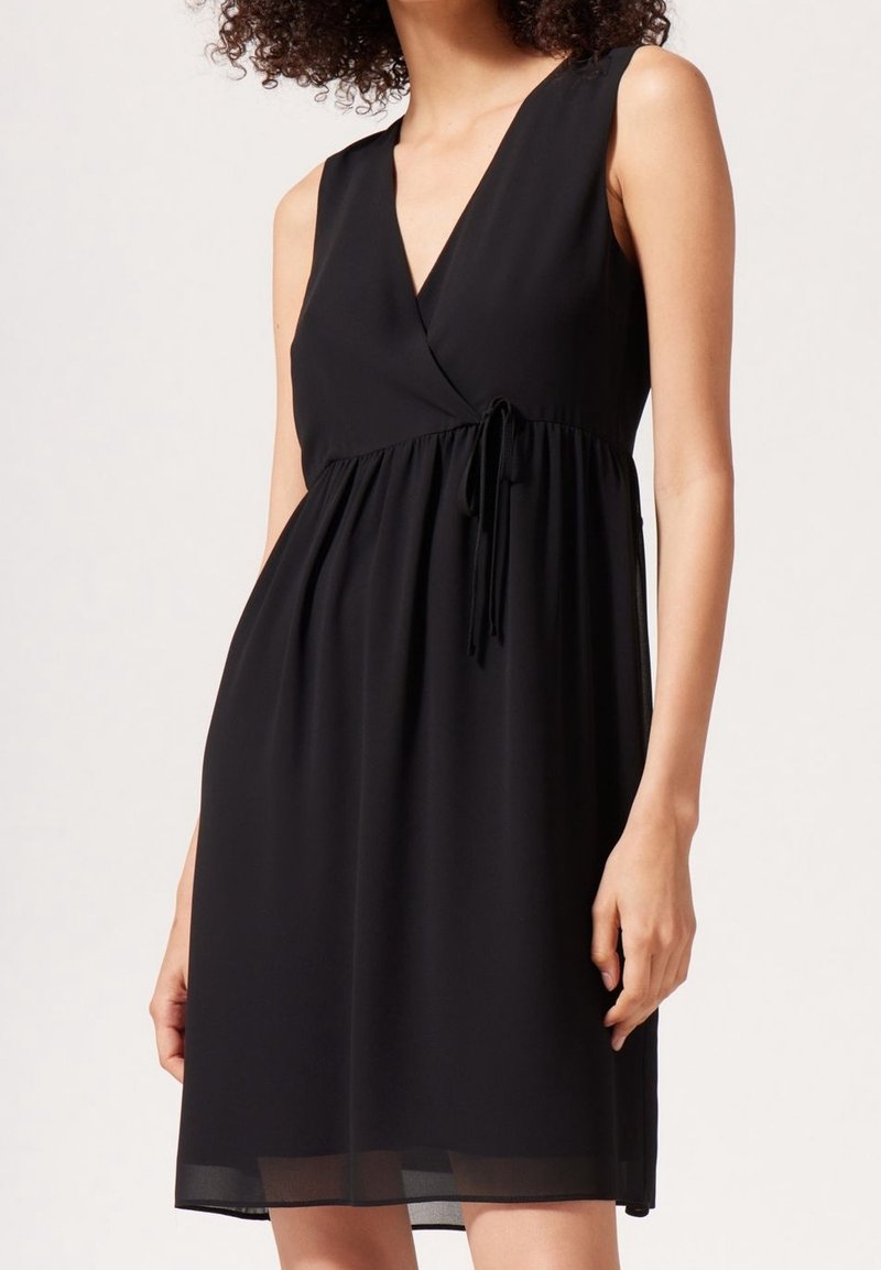 Robe noire sans manches en tissu léger, avec un col en V, un design cache-cœur, une taille ajustable par cordon et une silhouette fluide avec un ourlet transparent.
