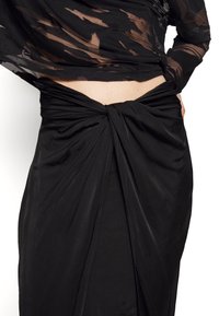 Vestido preto drapeado com cintura atada, feito de tecido leve com uma textura suave e brilho sutil. Parte superior translúcida com padrões abstratos.