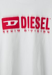T-shirt en coton blanc avec un logo rouge bien visible portant les inscriptions "DIESEL" et "DENIM DIVISION". Texture lisse et détails d'impression clairs.