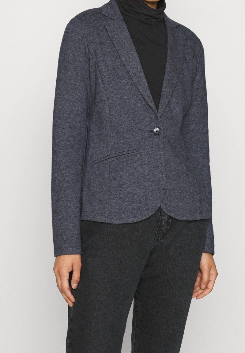 Námořnický modrý blazer vyrobený z měkké tkaniny s přiléhavým střihem, na jedno knoflíkové zapínání a se dvěma předními kapsami. Model má pod ním černý rolák.