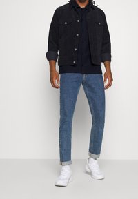 Chaqueta de pana azul oscuro con botones plateados, llevada sobre una camisa navy, combinada con jeans de mezclilla azul claro enrollados y zapatillas blancas.