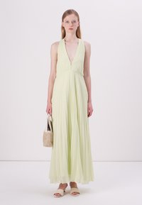 Abito maxi senza maniche verde chiaro con scollatura a V profonda e design plissettato, abbinato a una piccola borsa testurizzata e sandali semplici.