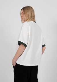 Plavokosa osoba u širokoj bijeloj majici s tamnozelenim rubom na rukavima i crnim hlačama, stojeći s rukama u džepovima.