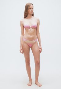 Γυναίκα ξυπόλυτη, ντυμένη με ροζ και κίτρινο μπικίνι ombre με λεπτές τιράντες, κοιτάζει στο πλάι πάνω σε απλό λευκό φόντο.