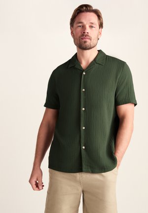 Man with light brown hair and beard wearing a short-sleeve dark green button-up shirt and beige pants, standing with one hand in pocket.