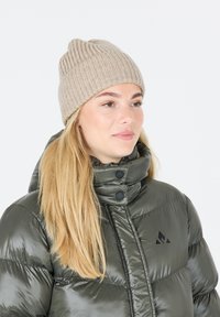 Beige ribbet strikket hue med en rund top, b�året af en person i en mørkegrøn pufferjakke med høj krave og sorte detaljer.