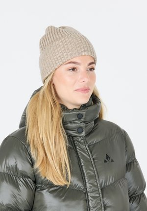 Beige ribbad stickad mössa med rundad topp, bärs av en person i en mörkgrön pufferjacka med hög krage och svarta detaljer.