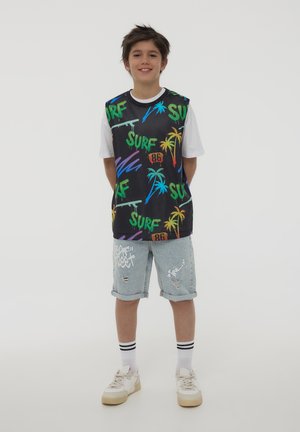 Canottiera nera in rete con testo "SURF" colorato e stampe di palme, abbinata a pantaloni shorts in denim strappati e sneakers bianche.