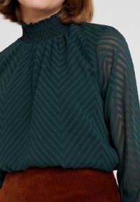 Blouse teal avec un col smocké haut et des manches longues transparentes, présentant un motif chevron texturé. Associée à une jupe en velours côtelé couleur rouille.