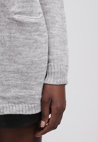 Šedý pletený svetr s žebrovanými manžetami a přední kapsou, s texturovaným povrchem a mírně volným střihem. Ruka modelky spočívá na lemu.