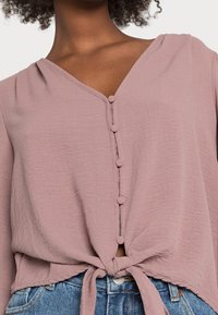 Femme portant une blouse boutonnée rose poussiéreux nouée à la taille, associée à un jean en denim bleu, montrant le haut du torse et le cou.