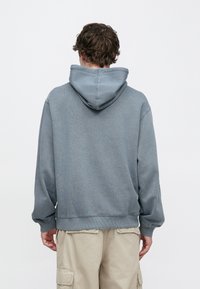 Szara bluza z kapturem o fakturowanym wykończeniu, z kapturem ze sznurkiem i ściągaczami na rękawach. Noszona z jasnobeżowymi spodniami typu cargo.