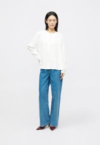 Bílá halenka s dlouhým rukávem a výstřihem ve tvaru klíčové dírky, v kombinaci s modrými širokými džíny a burgundskými botami na podpatku s špičatou špičkou. Hladká látka, volný střih.