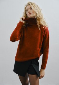 Rode, gebreide coltrui met geribbelde manchetten en zoom. Gedragen met zwarte wikkelshorts. Zachte textuur, relaxte pasvorm en casual ontwerp.