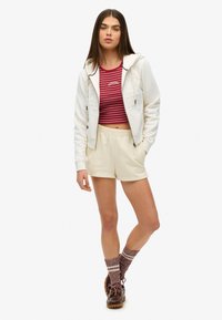 Witte zip-up hoodie met reliëftekst, bordeauxrode en witte gestreepte crop top, crème korte broek, bruine schoenen en ribbelige bordeauxrode sokken met witte strepen.