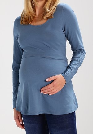 Persona incinta che indossa una maglia blu a maniche lunghe e jeans, tenendo delicatamente la pancia con una mano.