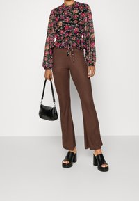 Blusa floreale a maniche lunghe con vita cintrata, abbinata a pantaloni a campana marroni a costine e sandali neri con tacco a blocco e dita scoperte, mentre tiene una borsa nera.