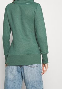Felpa verde realizzata in tessuto morbido, con colletto alto, polsini a coste e orlo leggermente più lungo. Indossata con jeans baggy blu chiaro.