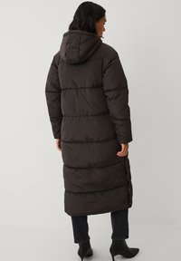Lange, donkerbruine pufferjas met een capuchon, voorzien van een gestikt ontwerp en zijknopen. Gedragen over donkere jeans en enkellaarzen.