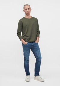 Homme portant une chemise olive à manches longues, un jean bleu et des baskets blanches, debout les mains dans les poches sur un fond clair uni.