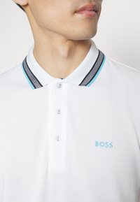 Homme portant un polo blanc avec un col rayé bleu et noir et trois boutons, brodé d'un logo bleu "BOSS" sur la poitrine.