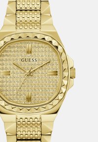 Reloj dorado con una esfera cuadrada que presenta una esfera texturizada adornada con cristales y una correa con tachuelas para un detalle adicional.