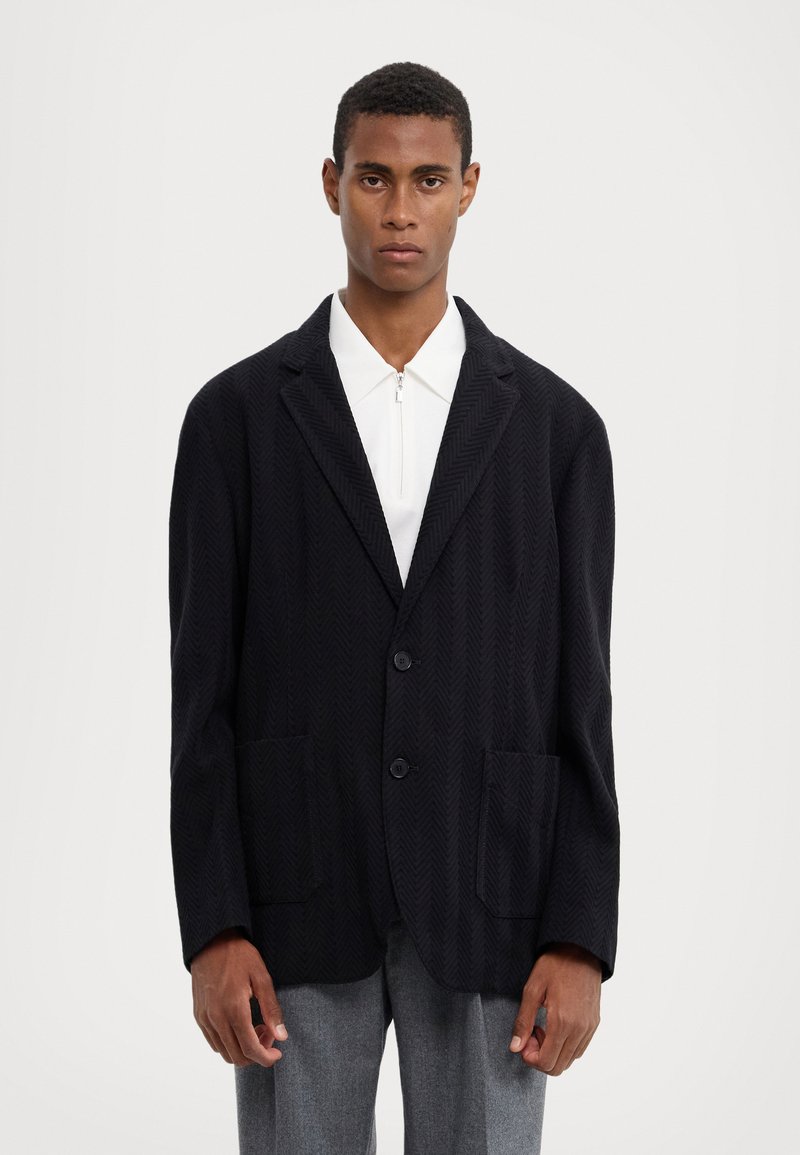 Blazer noir en chevrons avec deux poches avant et deux boutons. Doté d'un col cranté et d'un tissu texturé, associé à une chemise blanche.