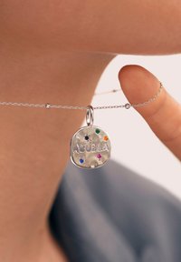 Pendente in argento con "ABUELA" inciso, adornato con diversi gemstone colorati. Attaccato a una catena delicata, tenuto da un dito.