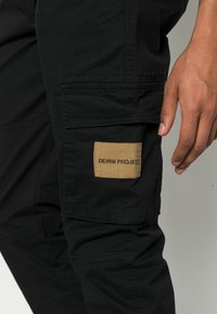 Čierne cargo nohavice s textúrovaným povrchom, s béžovou nášivkou s nápisom "DENIM PROJECT" a viacerými vreckami pre praktické využitie.
