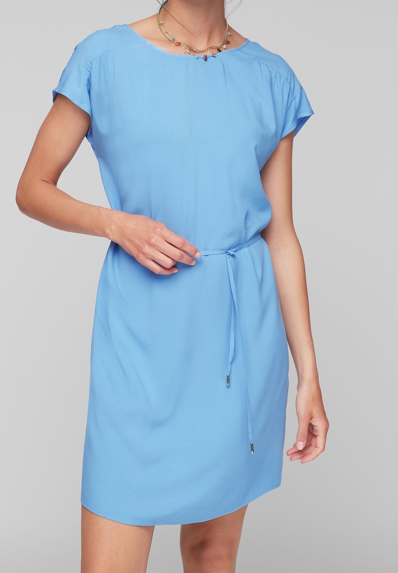 Femme portant une robe bleu clair à manches courtes avec une fine ceinture à la taille et des colliers en chaîne dorée superposés, debout devant un fond gris uni.