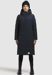 Cappotto lungo e voluminoso di un blu navy scuro con cappuccio, tasche con zip e silhouette dritta. Presenta una texture trapuntata e un dettaglio del logo sull'orlo.