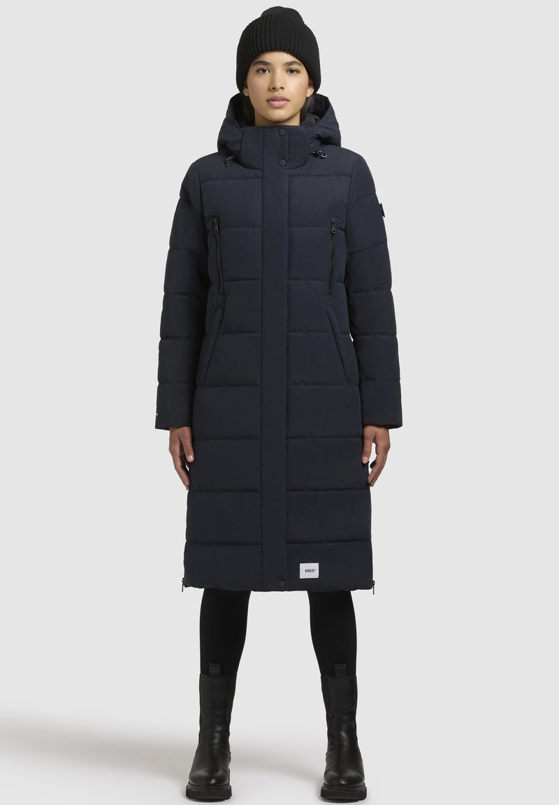 Cappotto lungo e voluminoso di un blu navy scuro con cappuccio, tasche con zip e silhouette dritta. Presenta una texture trapuntata e un dettaglio del logo sull'orlo.