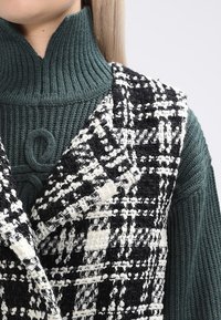 Kamizelka z tweedu w monochromatyczną kratę, noszona na prążkowanym zielonym swetrze z wysokim kołnierzem i teksturowaną tkaniną.