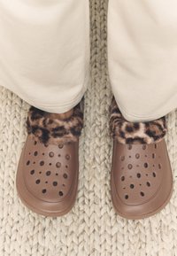 Brown synthetic clogs featuring a leopard-print faux fur cuff, with ventilation holes and a smooth, rounded design, set on a textured surface.