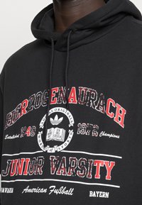 Un sweat à capuche noir avec du texte rouge et blanc, affichant "HERZOGENAURACH," "JUNIOR VARSITY," et un logo Adidas Originals avec des mots sur le thème du sport.