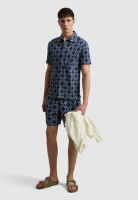 Chemise à manches courtes bleu marine et short assorti avec un motif géométrique blanc. Le modèle tient une chemise crème claire et porte des sandales à glisser marron.