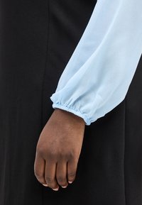 Σκούρο δέρμα χεριού ακουμπά σε μαύρο ύφασμα, φορώντας ένα ανοιχτό μπλε μακρυμάνικο με ελαστική μανσέτα που μαζεύει στον καρπό.