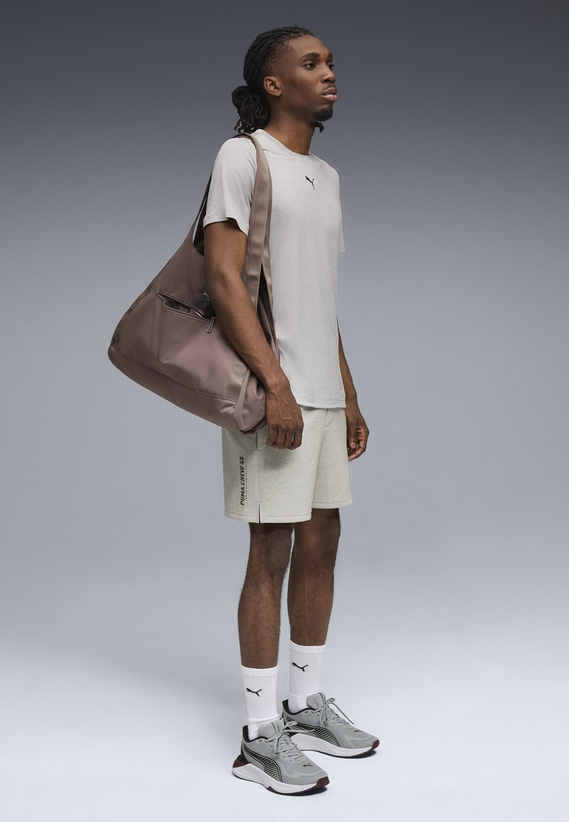 Graues T-Shirt mit Logo, hellfarbene Shorts und graue Turnschuhe mit schwarzen Akzenten. Braune Umhängetasche mit einem Reißverschlussfach.