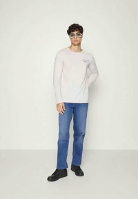 Camisa branca de manga longa com logótipo, jeans azuis e sapatos pretos. Modelo a usar óculos de sol verdes claros, de pé contra um fundo neutro.