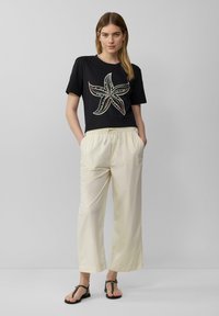 Zwarte crop t-shirt met een kleurrijke zeester ontwerpen; crème wijde broek; zwarte platte sandalen met een band, handen in de zakken.