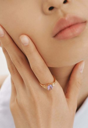 Anillo de oro con una gema púrpura central, con delicados acentos en forma de alas. Mano posicionada cerca de un rostro con piel suave.