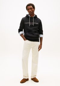 Sudadera con capucha azul marino con logo de texto blanco, bolsillo gris, combinada con pantalones crema y zapatos de ante marrón. Cuenta con cordones ajustables y puños en capas.