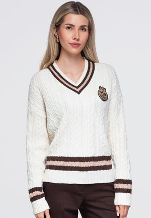 Une femme porte un pull blanc en tricot torsadé, orné de bordures à rayures marron et d’un écusson, assorti à un pantalon foncé, sur un fond uni.