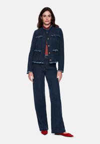 Dunkelblaue Jeansjacke mit ausgefransten Kanten und goldenen Knöpfen, kombiniert mit passenden Weitbein-Hosen. Rote flache Schuhe und ein gemustertes Oberteil sind sichtbar.