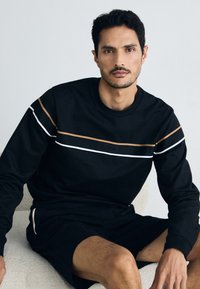 Svart langermet genser med horisontale striper i hvitt og gull, kombinert med matchende svarte shorts. Myk tekstur, avslappet passform.