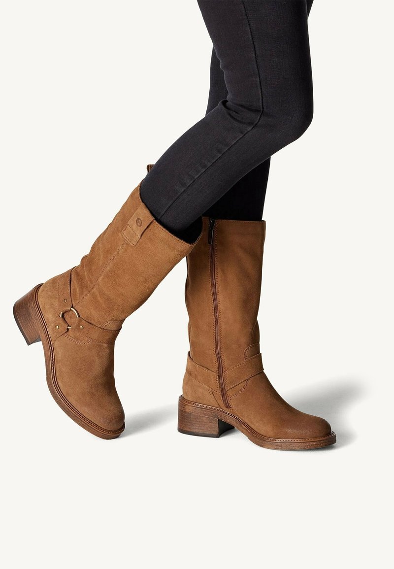 Jambes en pantalon noir portant des bottes en suède marron mi-mollet avec des talons blocs bas et une fermeture éclair latérale, présentées sur un fond clair uni.