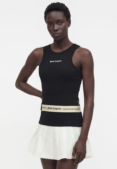 Γυναίκα με μαύρο αμάνικο κορμάκι και λευκή πλισέ φούστα, με το κείμενο «palm angels» στο κορμάκι και αντίστοιχη ζώνη μέσης.